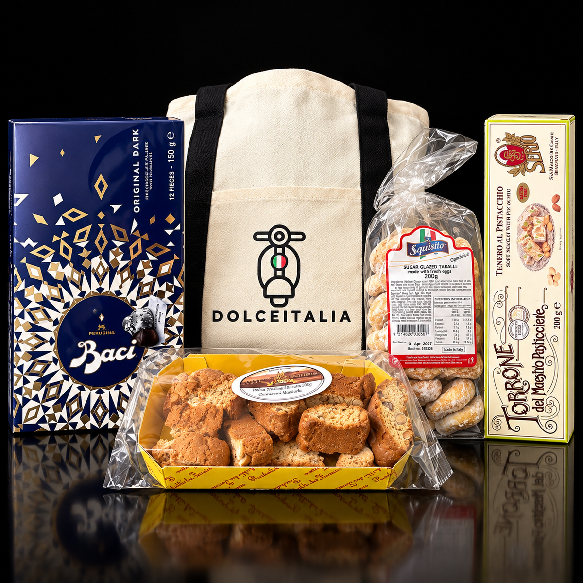 A Dolceitalia gift basket featuring a cream canvas tote bag with a black handle and logo, a blue and gold box of Baci Originali Dough, a bag of biscotti, and a box of Torrone Noto Pasticcino, arranged on a reflective black surface.